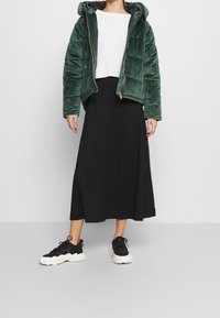 Grön vadderad jacka med huva och dragkedja, bärs över en vit profilerad tröja och svart midiklänning. Svarta sneakers med vita sulor.