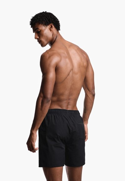 Μυώδης άνδρας με σκουρόχρωμο δέρμα και κοντά σγουρά μαλλιά, φοράει μαύρα μαγιό και στέκεται κοιτώντας πάνω από τον ώμο του.