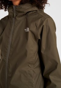 Oliivinvihreä vedenpitävä takki, jossa on vetskari etuosassa, säädettävä huppu ja The North Face -logo rinnassa. Pehmeä, matta pinta.