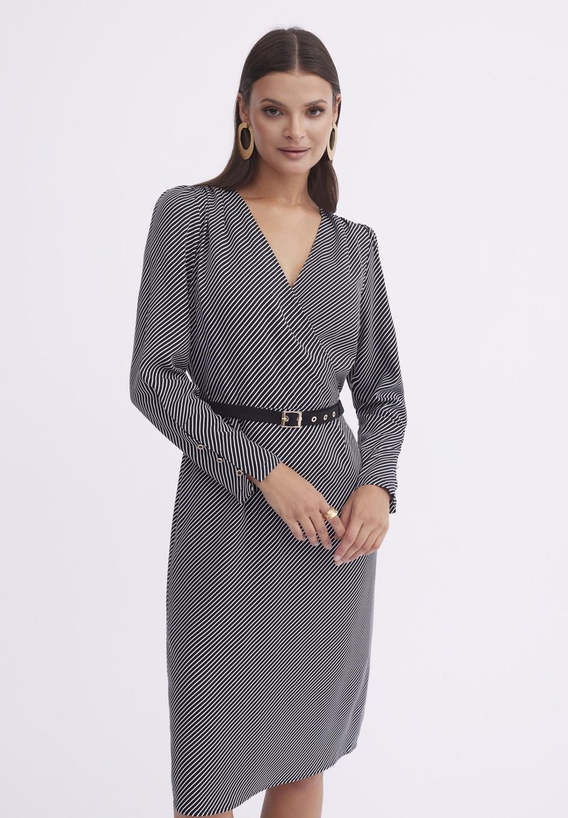 Czarna i biała sukienka w paski z fasonem kopertowym, długimi rękawami i dopasowaną sylwetką. Udekorowana czarnym paskiem i guzikami.