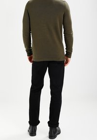 Maglietta a maniche lunghe in maglia verde oliva, slim fit, abbinata a pantaloni neri; texture liscia, dettagli minimi e stivaletti neri alla caviglia.