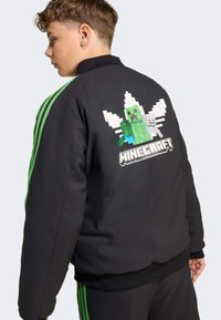Svart bomberjacka med gröna detaljer, som har en pixeliserad Minecraft-creeper och karaktärer på ryggen, ribbad krage och muddar.
