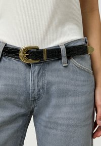 Cinturón negro con estampado de cocodrilo y hebilla en tono dorado, con un diseño acanalado y acentos en punta, usado sobre unos jeans de mezclilla azul claros.