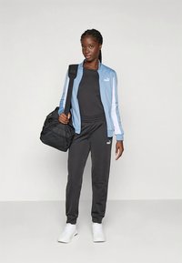 Blå jacka med dragkedja och vita ränder, svart figursydd tröja, svarta mjukisbyxor och en svart sportbag. Vita sneakers kompletterar outfiten.