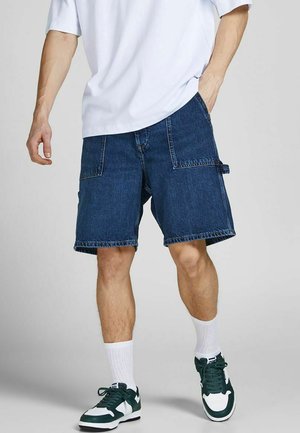 Dunkelblaue Denim-Cargoshorts mit Seitentaschen und weiter Passform, kombiniert mit weißem T-Shirt und grünen Turnschuhen.