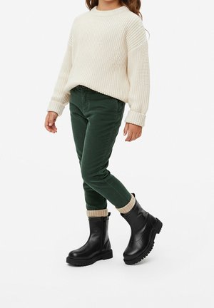 Dziecko ubrane w kremowy sweter z dzianiny, ciemnozielone spodnie i czarne botki z beżowymi dzianinowymi mankietami na białym tle.