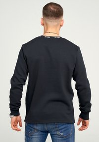 Hombre con cabello corto que lleva un sudadera negra con "JCK & JNS" en el cuello, jeans azules desgastados y un reloj de pulsera plateado, de espaldas.