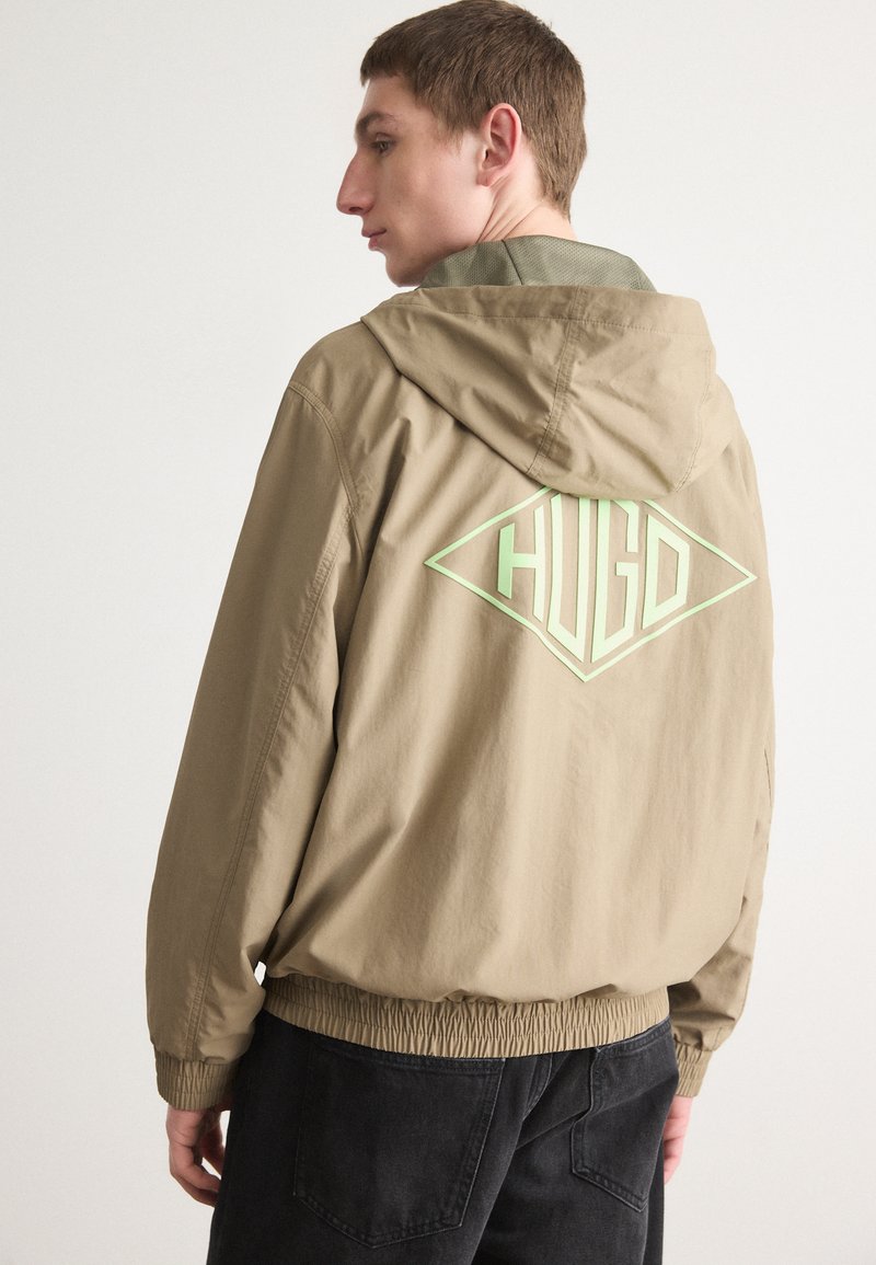 Beige hette jakke med ribbede mansjetter, og med en stor grønn logo på baksiden. Glatt stoff med en avslappet passform og elastisk midje.