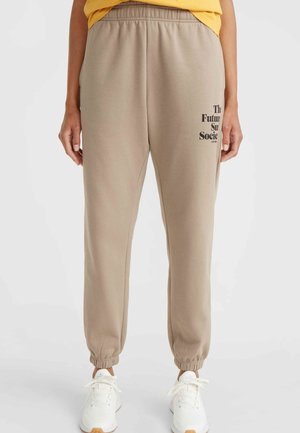 Pantalon de jogging beige avec taille et poignets élastiques, texte noir sur la cuisse gauche, porté avec des baskets blanches et un haut jaune.