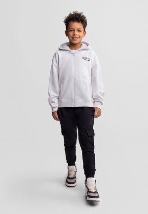 Uśmiechający się chłopiec w białej bluzie z zamkiem, czarnych spodniach typu cargo i czarno-białych trampkach, stojący na tle jednolitego szarego tła.