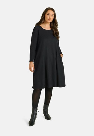 Mujer con un vestido negro suelto de manga larga, medias negras y botines negros, de pie frente a un fondo blanco.