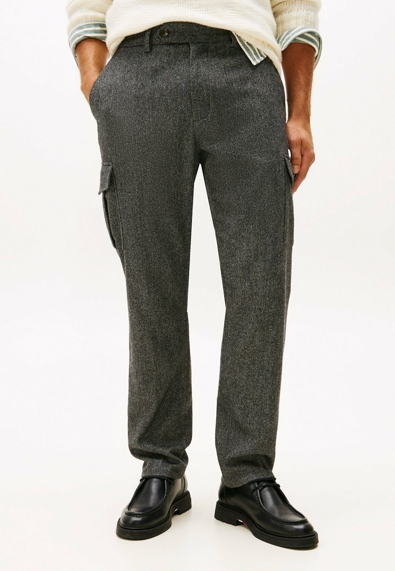 Hombre que lleva pantalones cargo en gris de espiga con bolsillos laterales, zapatos negros de piel con cordones, un suéter color crema y puños de camisa a rayas visibles.