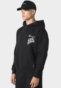 Zwarte hoodie van katoen, met een voorkantzak, geborduurd logo in wit en roze, en ribbels aan de mouwen. Het model draagt ook een zwarte pet.