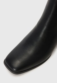 Botín de cuero negro hasta el tobillo con punta cuadrada, textura suave y un ligero tacón, diseñado con líneas minimalistas y sin herrajes visibles.