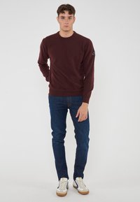 Maglione bordeaux con maniche lunghe, collo rotondo e una toppa con logo sulla spalla sinistra. Indossato con jeans blu scuro e sneaker bianche.