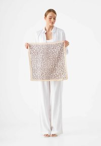 Foulard en soie à imprimé léopard avec une bordure beige, de forme carrée, tenu par une personne en tenue blanche devant un fond clair.