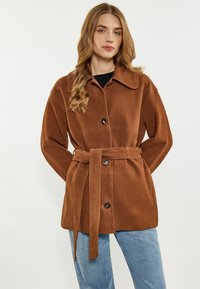 Giacca in suede marrone con collo a camicia, chiusura con bottoni e vita con cintura. Presenta una vestibilità rilassata e due tasche frontali.