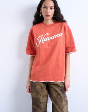 Kvinde med langt mørkt hår iført koralorange oversized T-shirt med teksten "Havana" og mønstrede brune bukser mod en ensfarvet hvid baggrund.