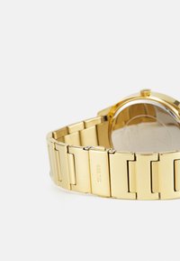 Gold leather watch with a linked band, round case, minimalistic design, and a small crown. Back shows a polished metal finish.