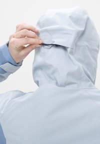 Veste imperméable bleu clair avec capuche, montrant une personne ajustant le cordon de serrage. Présente des coutures sans couture et une texture lisse.