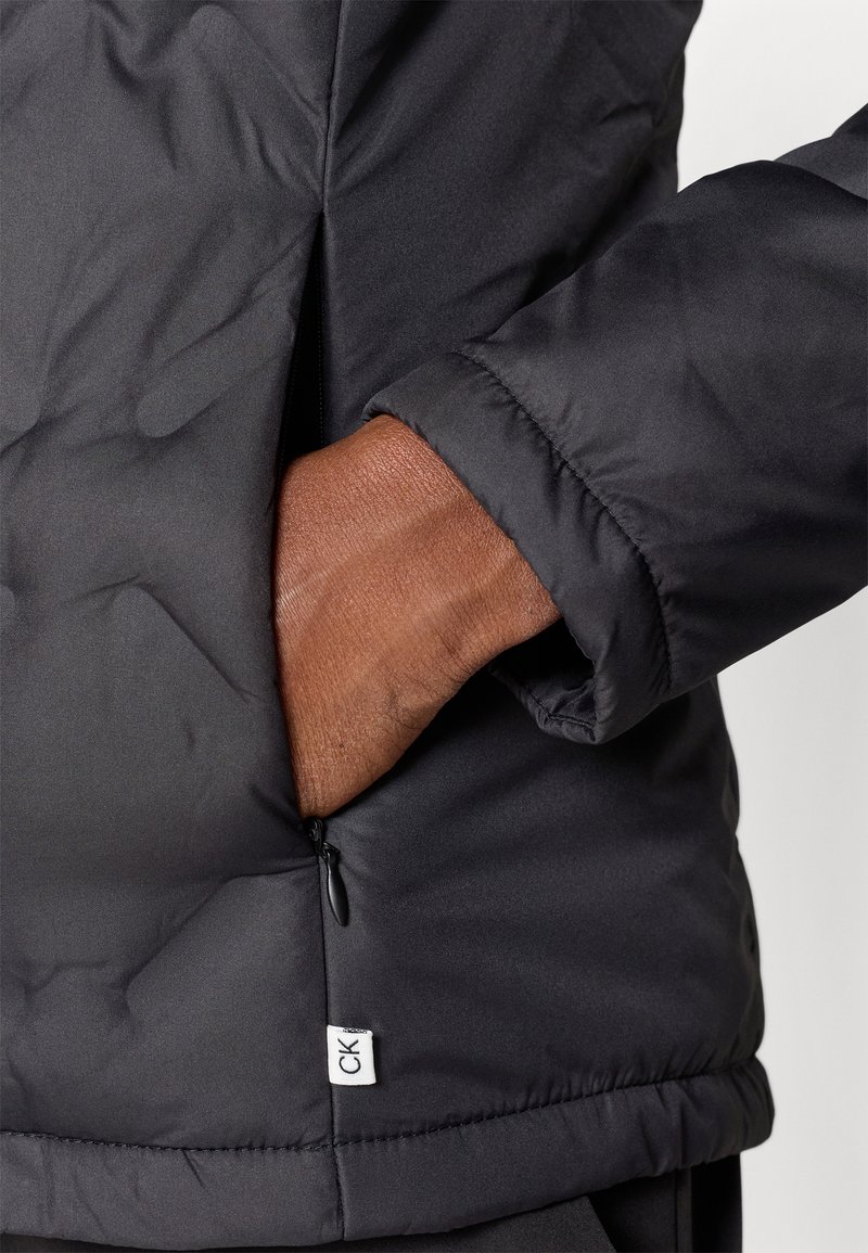 Giubbotto piumino nero con una texture liscia, tasca con zip e maniche arrotolate. Etichetta con il logo CK visibile all'orlo.