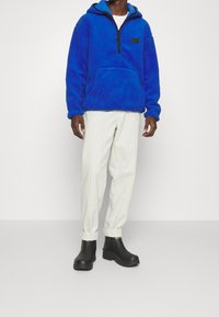 Felpa blu in pile con mezzo zip, tasca anteriore e toppa nera con logo; abbinata a pantaloni tapered bianchi a righe e stivali neri.