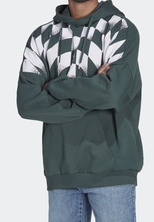 Hombre con sudadera con capucha verde oscuro con patrón de cuadros blancos en el pecho y vaqueros azul claro, brazos cruzados.