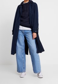Larga gabardina azul marino de tela brillante, combinada con una sudadera azul marino y una camiseta blanca. Unos vaqueros de tiro alto azul claro y zapatillas blancas completan el conjunto.