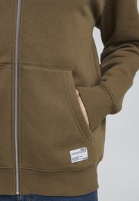 Braune Zip-Up-Hoodie aus weichem Stoff, mit seitlichen Taschen und einem Etikett am Saum. Die Bündchen und der Saum haben eine gerippte Textur.