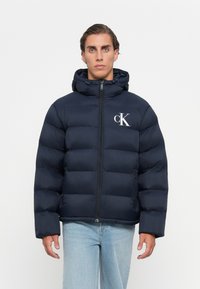 Man in een marineblauwe gewatteerde jas met capuchon en wit CK-logo, lichte blauwe spijkerbroek, staand tegen een effen witte achtergrond.