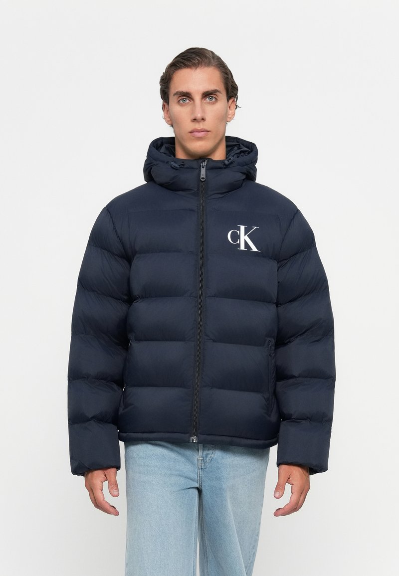 Man in een marineblauwe gewatteerde jas met capuchon en wit CK-logo, lichte blauwe spijkerbroek, staand tegen een effen witte achtergrond.