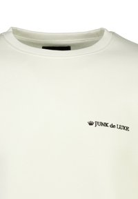 Sudadera blanca con cuello redondo, que presenta un pequeño logo negro "JUNK de LUXE" en el área del pecho izquierdo. Textura de tela suave, diseño minimalista.