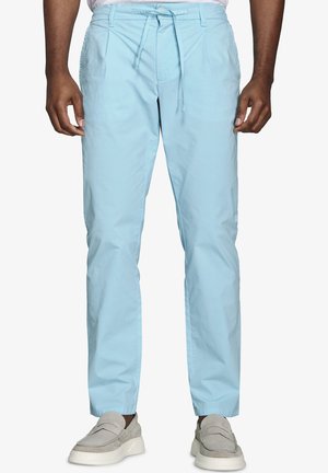 Mężczyzna w jasnoniebieskich spodniach z sznurkiem i beżowych butach wsuwanych, stojący na jednolitym białym tle.