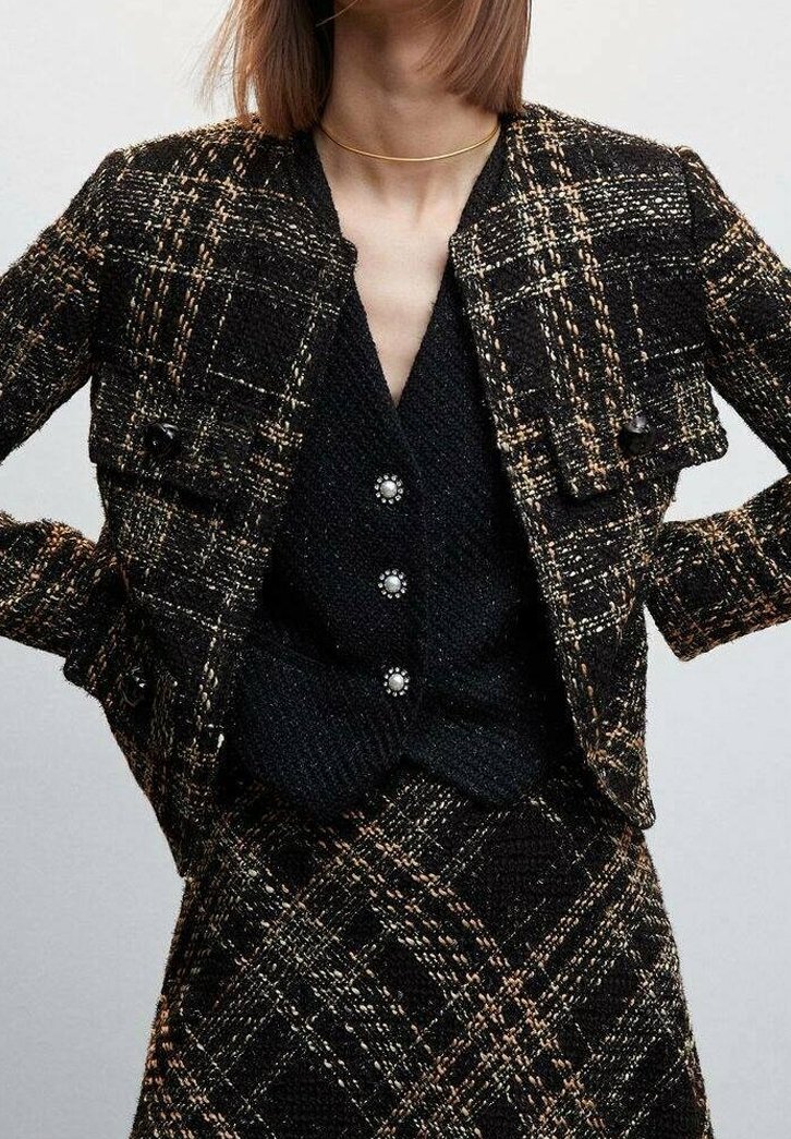 Femme portant un ensemble veste et jupe à carreaux texturés noir et marron sur un top noir boutonné avec des boutons en forme de perles et un fin collier ras-du-cou en or.