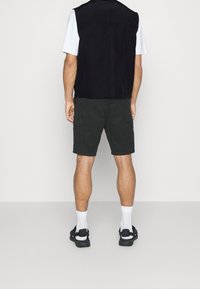 Mężczyzna ubrany w czarne spodenki cargo, czarną kamizelkę bez rękawów, białą koszulkę, białe skarpetki i czarne sandały, stojący tyłem na jednolitym tle.