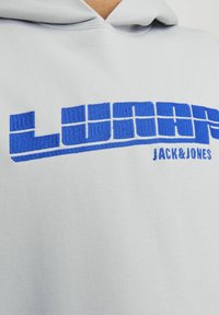 Sudadera gris claro con gran texto bordado en azul "LUNAR" con una tipografía estilizada, junto a "JACK & JONES" en texto más pequeño.