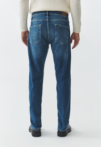 Jeans de mezclilla azul con corte recto, cinco bolsillos, costuras reforzadas y una etiqueta de cuero marrón en la cintura. Se llevan con zapatos negros.