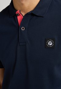 Mörkblå pikétröja med en rosa krage, försedd med två svarta knappar och en fyrkantig logotypapplikation på vänster bröst. Texturerad tyg.