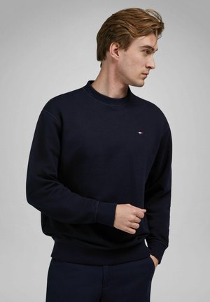 Junger Mann mit hellbraunem Haar, der einen marineblauen Pullover und passende Hose trägt, blickt nach rechts vor einem grauen Hintergrund.