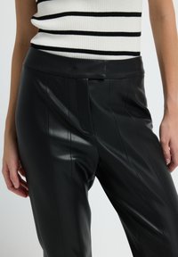 Zwarte faux leren broek met verticale naden, hoge taille en een gladde, glanzende textuur. Gestreepte top die gedeeltelijk zichtbaar is aan de bovenkant.