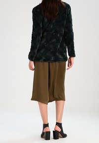 Tekstureret mørkegrøn faux pelsjakke med lange ærmer, kombineret med olivengrønne vidde-ben shorts og sorte åbne tå-hæle støvler.