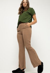Pantalones marrones ajustados con una textura suave, piernas acampanadas, bolsillos laterales y cintura alta, combinados con una camiseta de color verde oscuro y zapatillas negras.