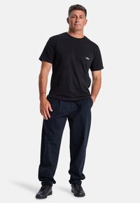 Zwarte katoenen T-shirt met een borstzak en geborduurd logo, gecombineerd met loszittende marineblauwe broek en zwarte schoenen.