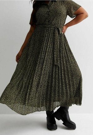 Femme portant une robe midi plissée à motifs verts avec une ceinture à la taille et des bottes noires épaisses, debout avec une main sur la hanche.