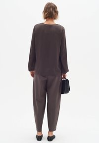 Brown long-sleeve shirt with a round neckline, paired with loose-fitting brown trousers and black shoes. Black bag held in one hand.