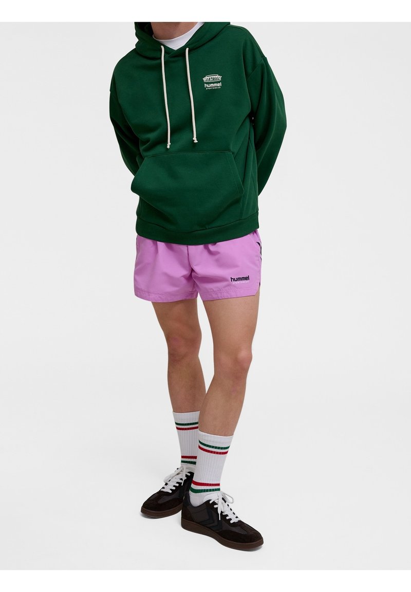 Personne portant un sweat à capuche vert foncé, un short de sport rose, des chaussettes blanches avec des rayures rouges et vertes, et des baskets noires, debout devant un fond uni.
