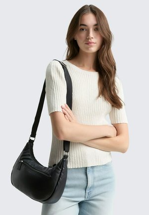 Schwarze synthetische Schultertasche mit einer geschwungenen Form, verstellbarem Riemen und einer vorderen Reißverschlusstasche, getragen über einem hellen gerippten Oberteil.