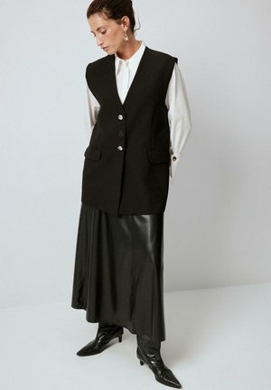 Black sleeveless vest with silver buttons, paired with a long black leather skirt and pointed black boots, set against a neutral background.