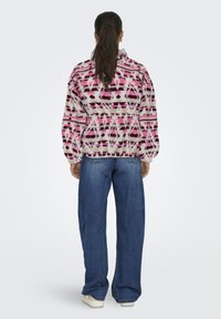 Frau mit langem, dunklem Pferdeschwanz, die eine rosa-weiß gemusterte Fleecejacke, blaue weite Jeans und weiße Sneakers trägt und von hinten steht.