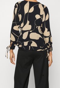 Schwarze und beige Bluse mit abstraktem Muster, langen Ärmeln mit Bindedetails, rüschigem Saum und glatter Textur. Kombiniert mit schwarzen Hosen.